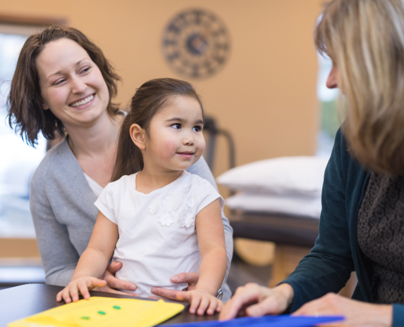 A young girl and her mother smile at an occupational therapist while sitting at a table