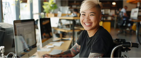 A young woman in a wheelchair sits at a computer and smiles at the camera